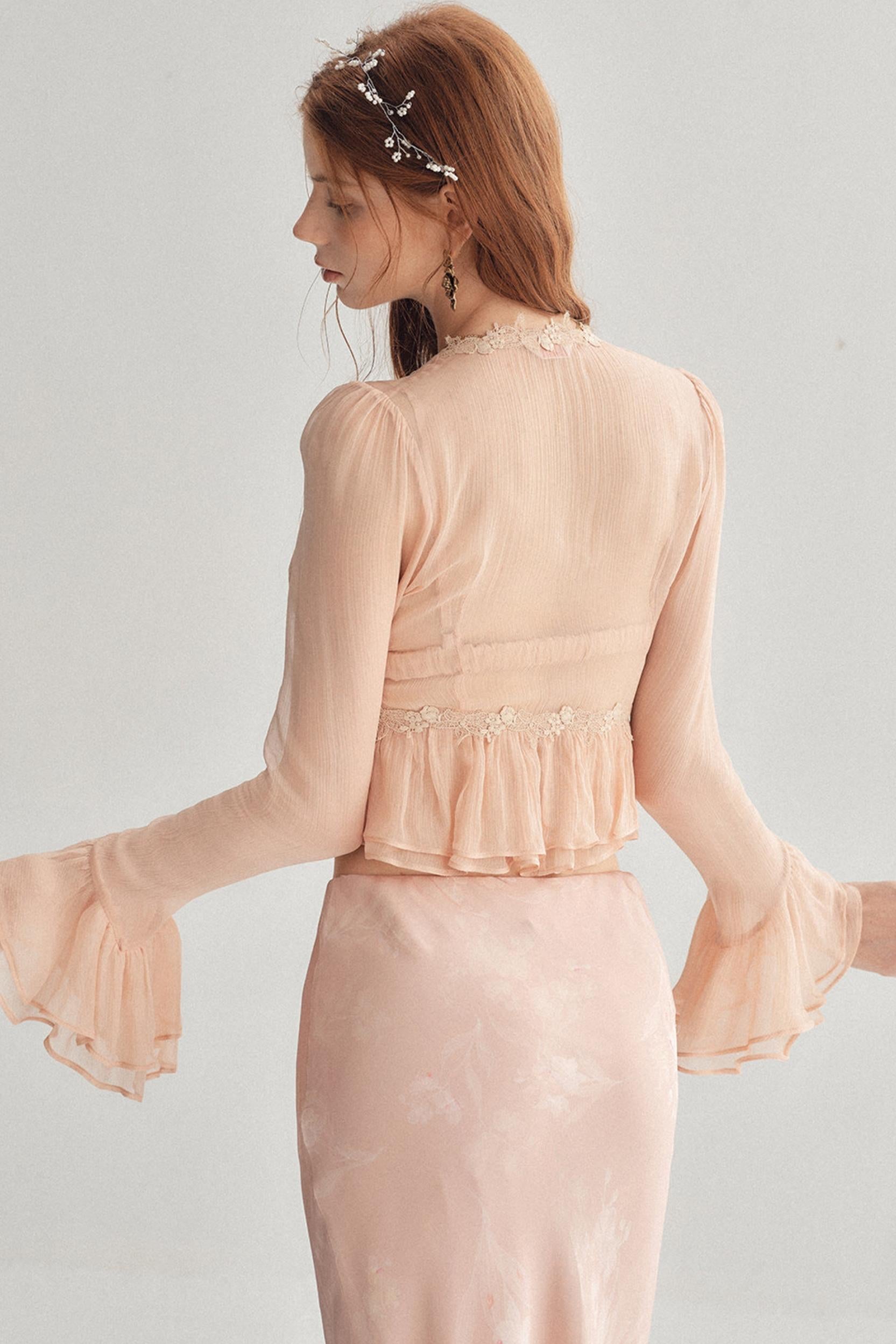 A woman shown from the back wearing a pale blush pink long-sleeve dress with a ruffled waistband and flared bell sleeves, against a light gray studio background.