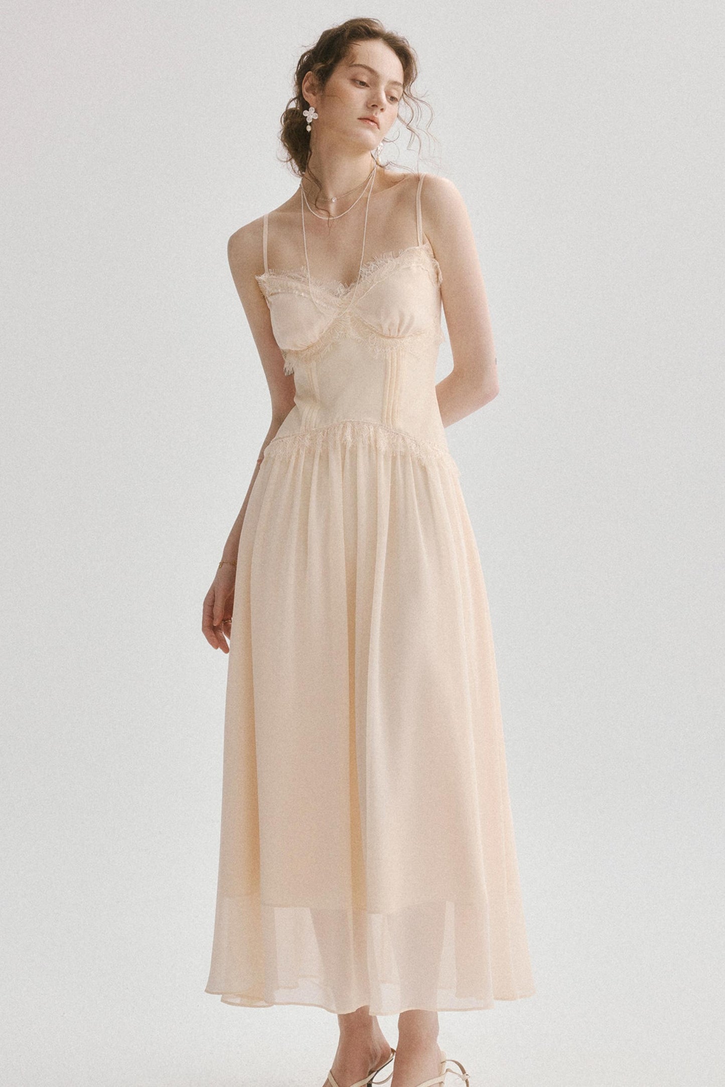 A woman modeling a light beige or cream midi dress with thin spaghetti straps, a sweetheart neckline, a structured corset-style bodice, and a flowing chiffon skirt, set against a plain light gray studio background.