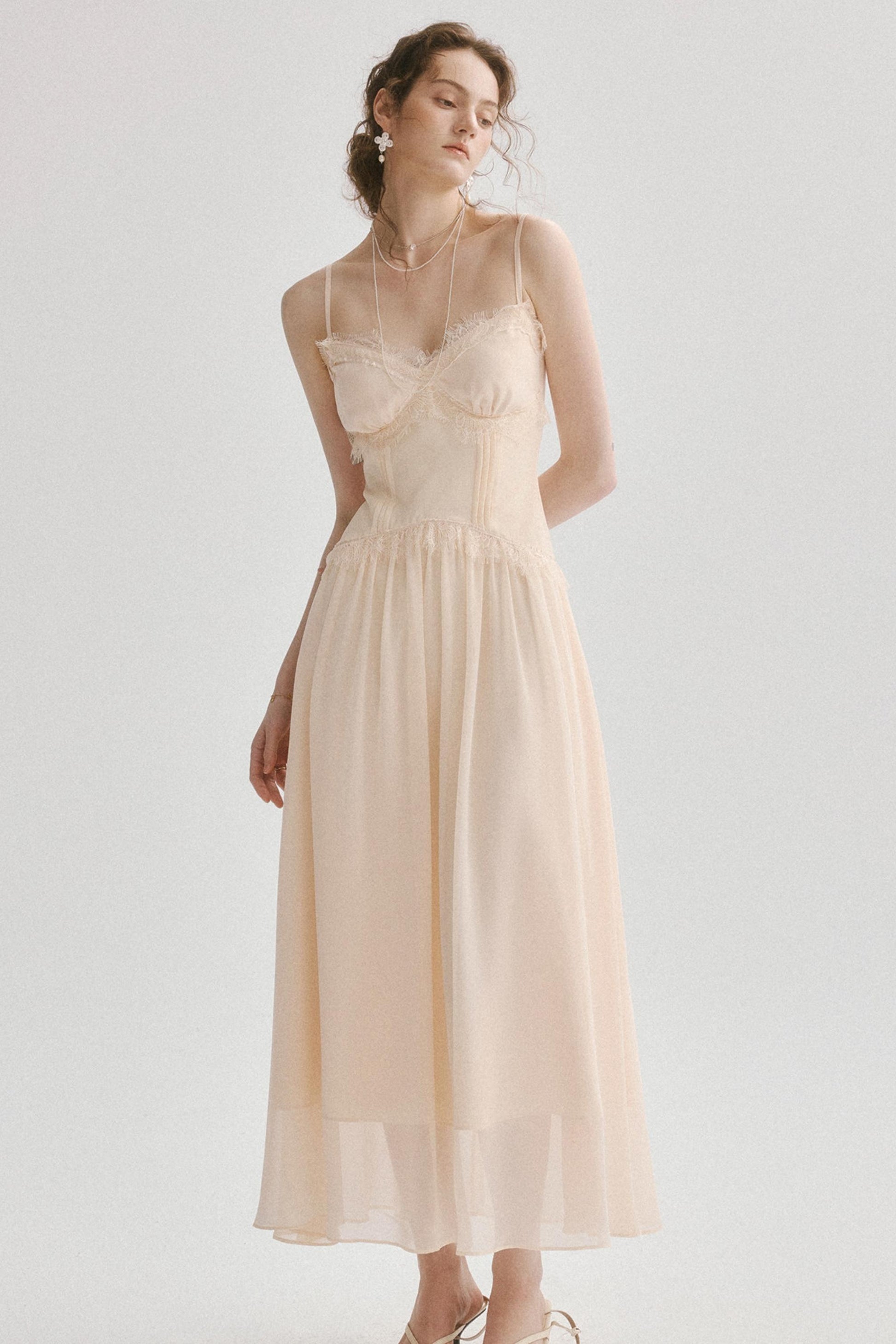 A woman modeling a light beige or cream midi dress with thin spaghetti straps, a sweetheart neckline, a structured corset-style bodice, and a flowing chiffon skirt, set against a plain light gray studio background.