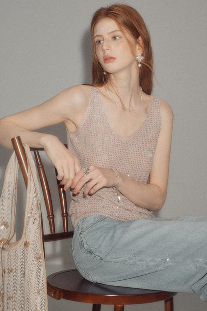 A woman with red hair sits on a wooden chair wearing a shimmering beige knit sleeveless top and light blue jeans, with a matching beige knit bag draped over the chair against a plain gray background.