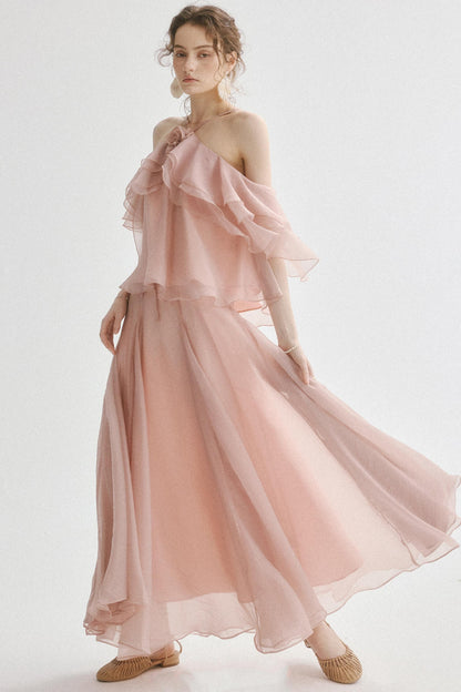 A woman modeling a flowing, light pink off-the-shoulder maxi dress with layered ruffle details and a full skirt, paired with beige flat shoes against a plain white studio background.