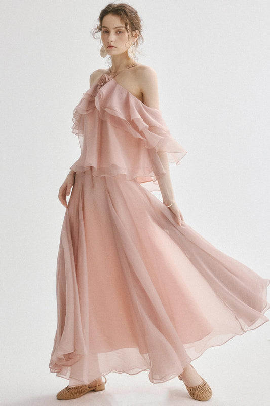 A woman modeling a flowing, light pink off-the-shoulder maxi dress with layered ruffle details and a full skirt, paired with beige flat shoes against a plain white studio background.