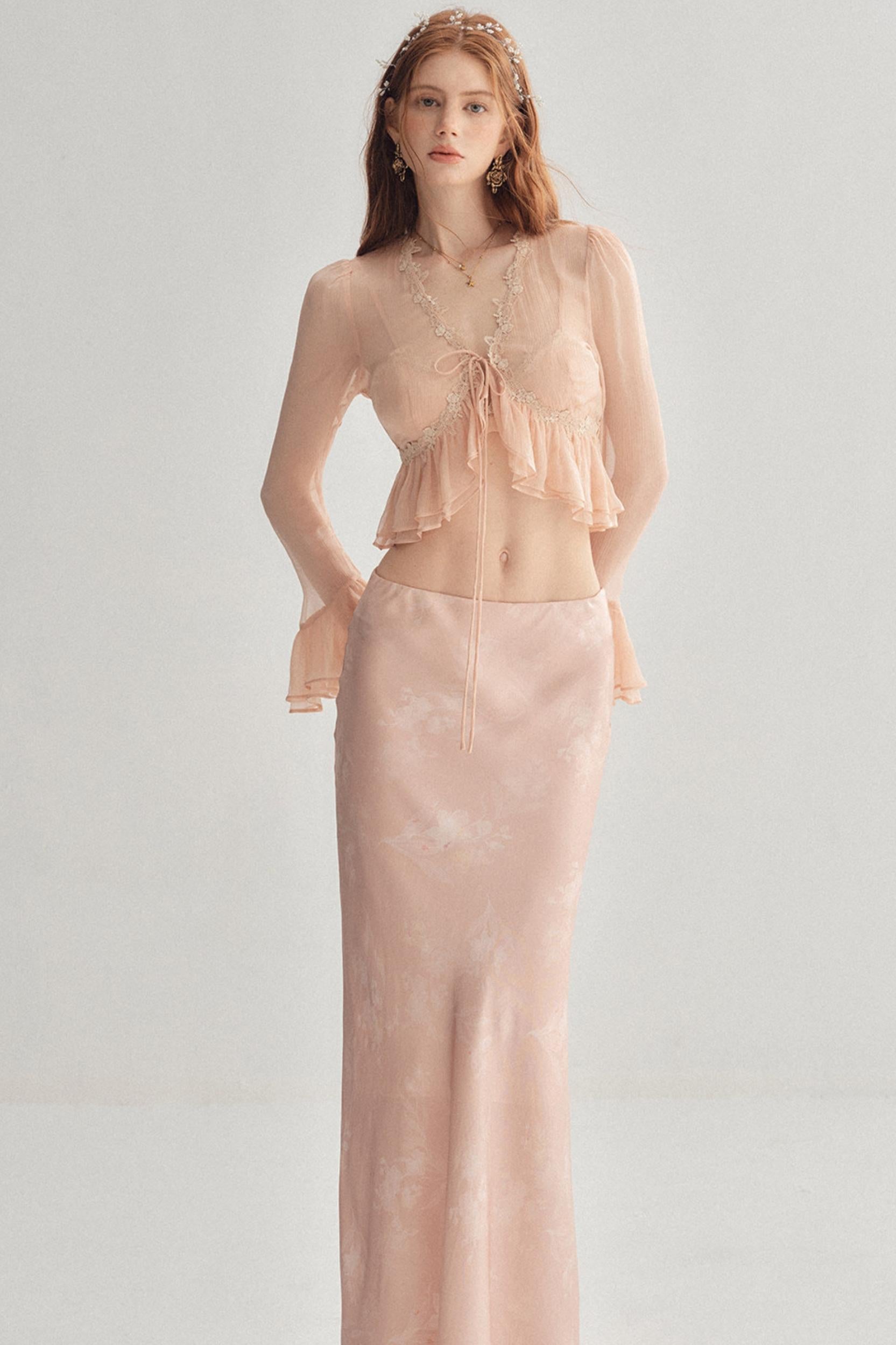 A woman modeling a two-piece peach pink set consisting of a sheer long-sleeve ruffled crop top with a tie-front detail and a matching satin-like long skirt, against a plain light gray studio background.