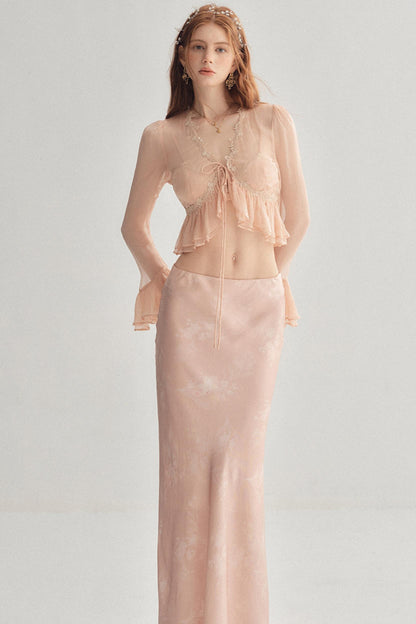 A woman modeling a two-piece peach pink set consisting of a sheer long-sleeve ruffled crop top with a tie-front detail and a matching satin-like long skirt, against a plain light gray studio background.