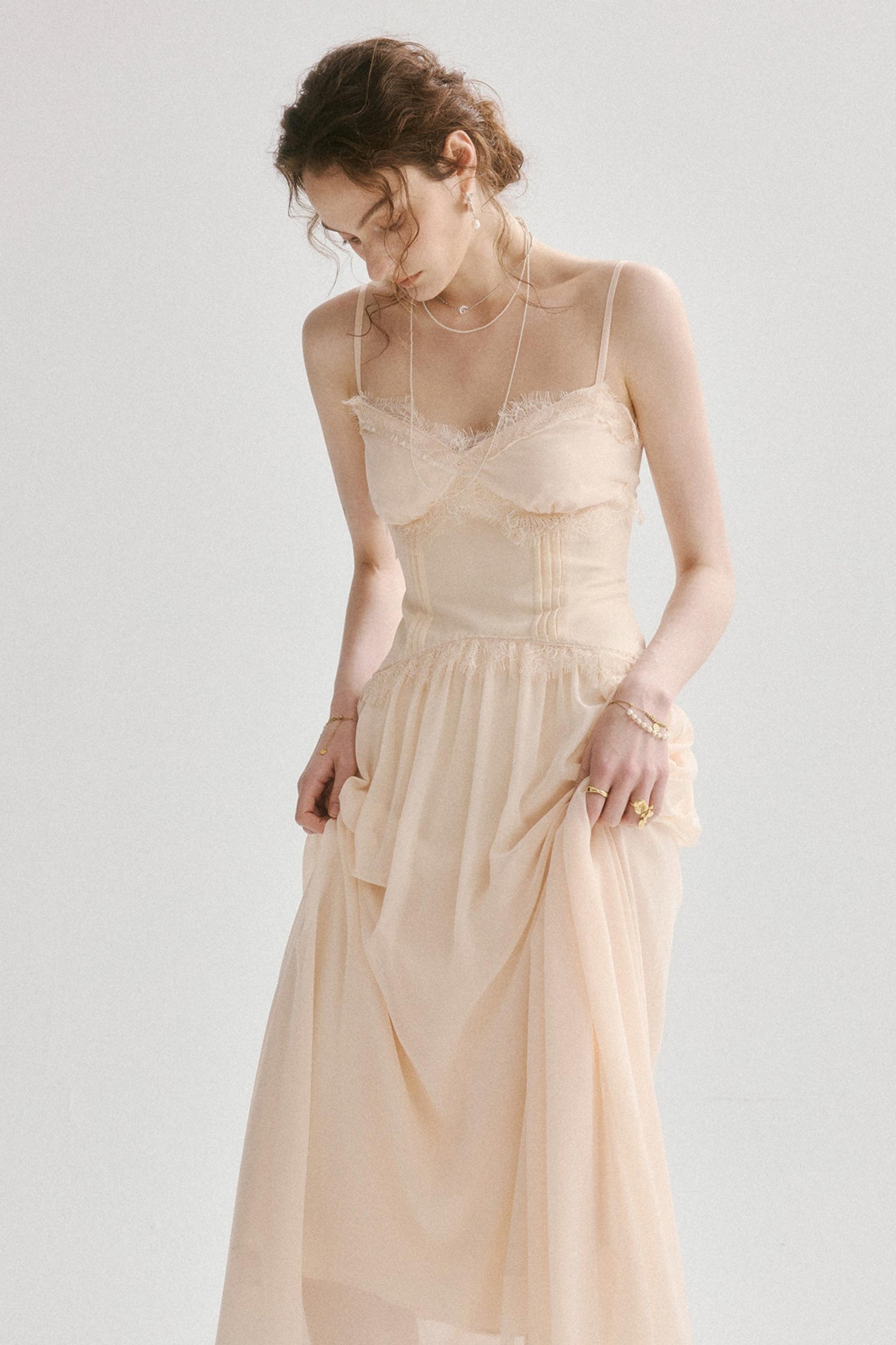 A woman wearing a light beige spaghetti-strap midi dress with a lace-trimmed sweetheart bodice and a flowing chiffon skirt, posed against a solid off-white background.