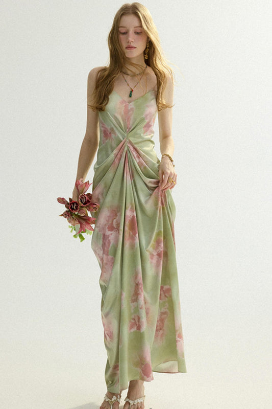 A woman modeling a sleeveless maxi dress with a sage green base and pink floral print, holding a small bouquet of pink flowers against a plain light background.