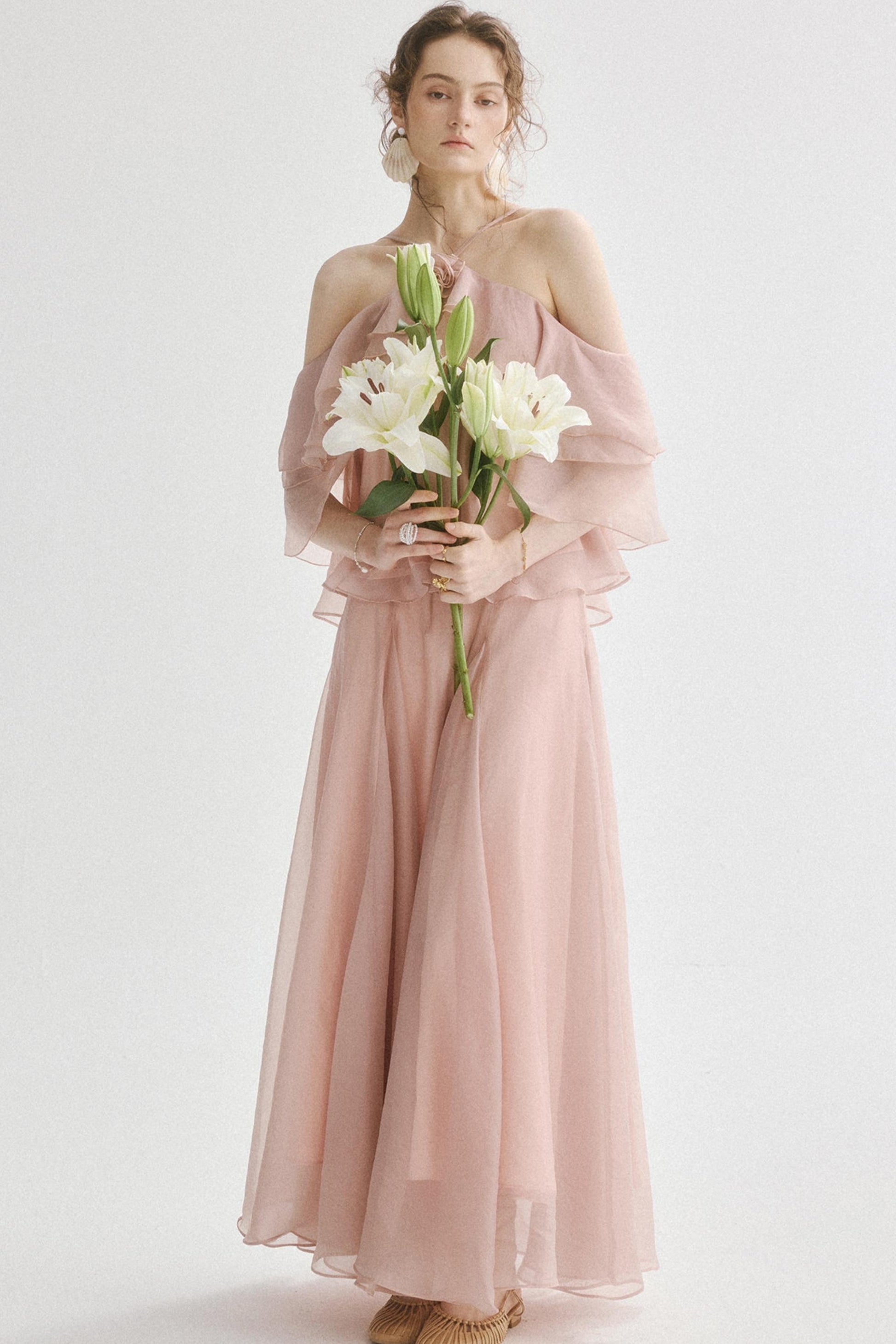A woman stands against a plain light background wearing a long blush pink off-the-shoulder tulle dress and holding a bouquet of white lilies.