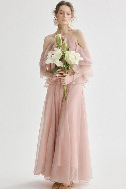 A woman stands against a plain light background wearing a long blush pink off-the-shoulder tulle dress and holding a bouquet of white lilies.