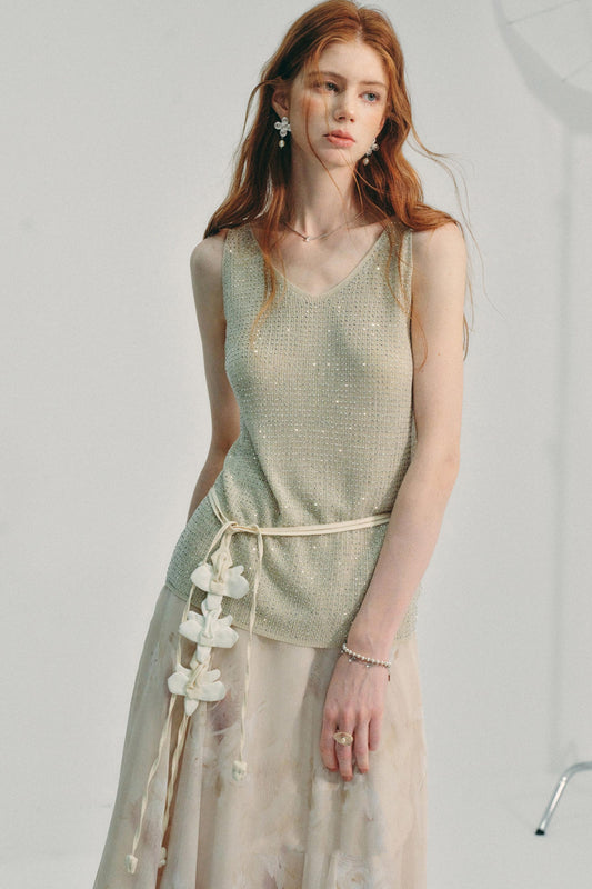 A woman modeling a sleeveless shimmering sage green top with a cream tie belt and a long sheer beige skirt, photographed against a plain light gray studio background.