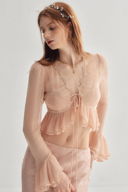 A woman wearing a sheer blush pink two-piece lingerie set with lace trim, a tie-front crop top, and matching ruffled bottoms, photographed against a plain light gray studio background.