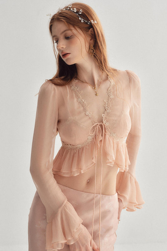 A woman wearing a sheer blush pink two-piece lingerie set with lace trim, a tie-front crop top, and matching ruffled bottoms, photographed against a plain light gray studio background.
