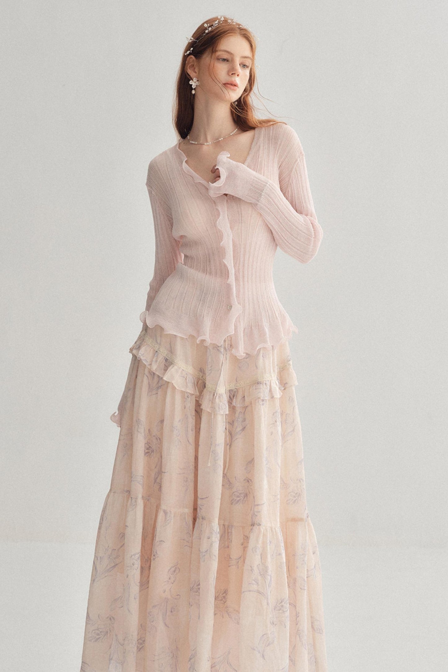 A woman modeling a long-sleeve light pink ribbed knit top with ruffle trim, paired with a matching long tiered skirt in a soft blush floral print, against a plain light gray studio background.