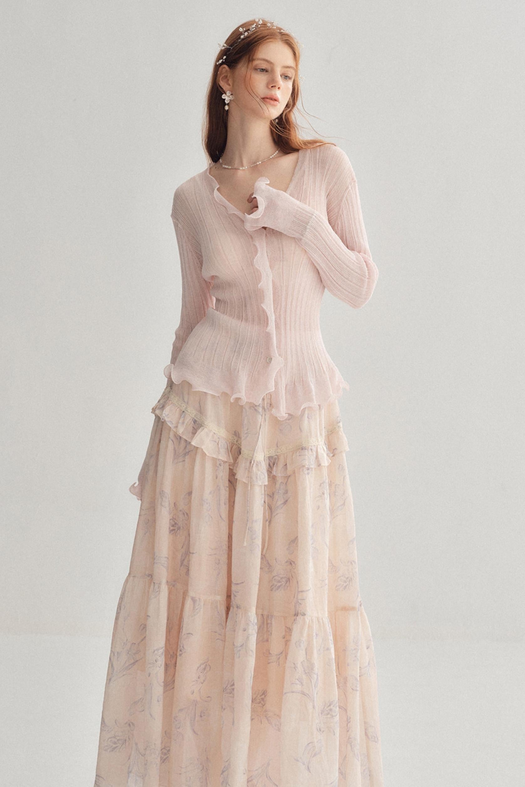 A woman modeling a long-sleeve light pink ribbed knit top with ruffle trim, paired with a matching long tiered skirt in a soft blush floral print, against a plain light gray studio background.