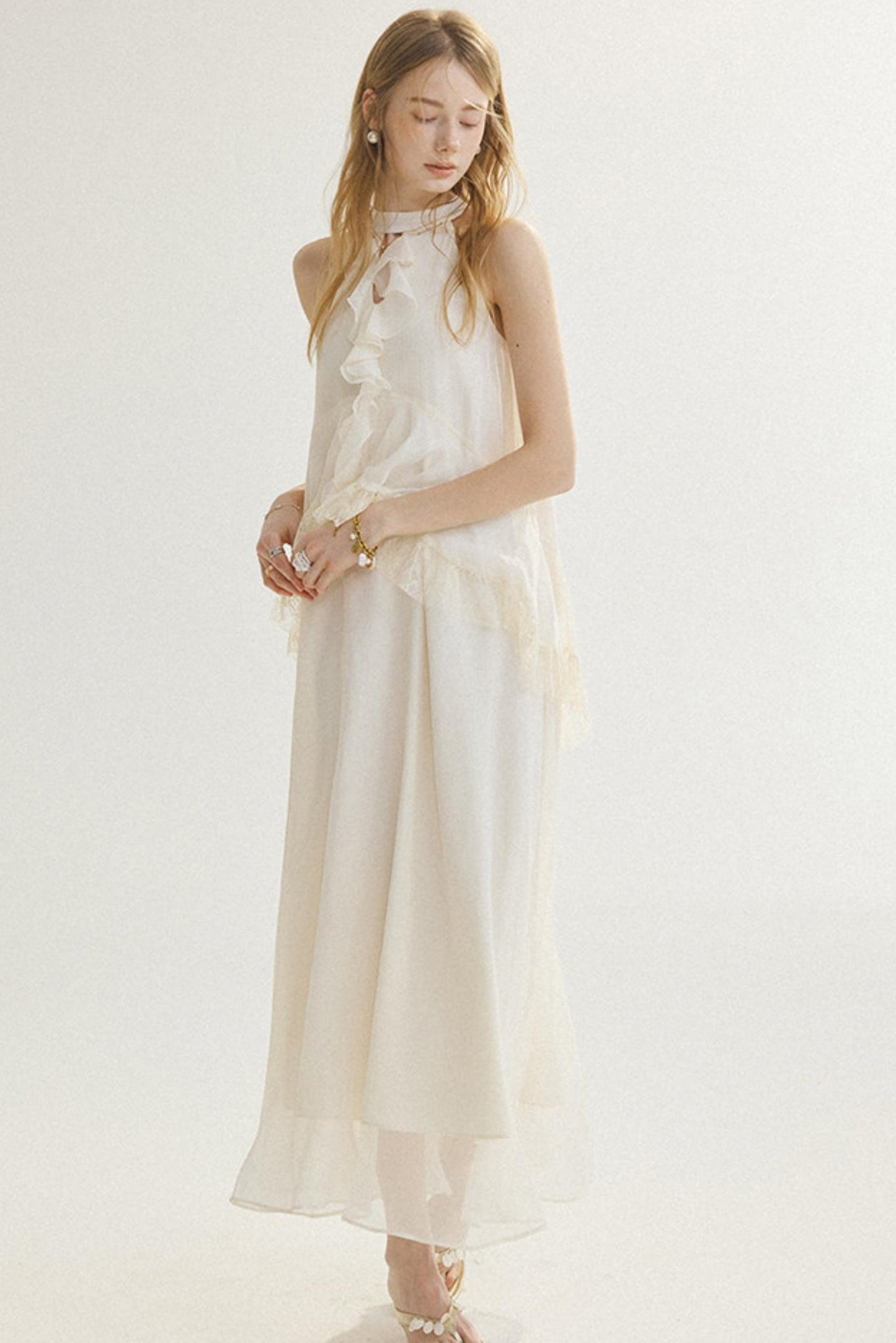 A woman modeling a sleeveless, high-neck, floor-length cream dress with layered ruffle details and sheer tulle overlay, set against a plain white studio background.