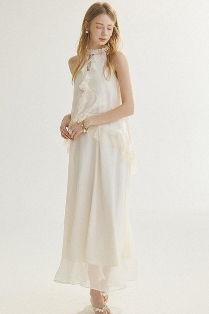 A woman modeling a sleeveless, high-neck, floor-length cream dress with layered ruffle details and sheer tulle overlay, set against a plain white studio background.