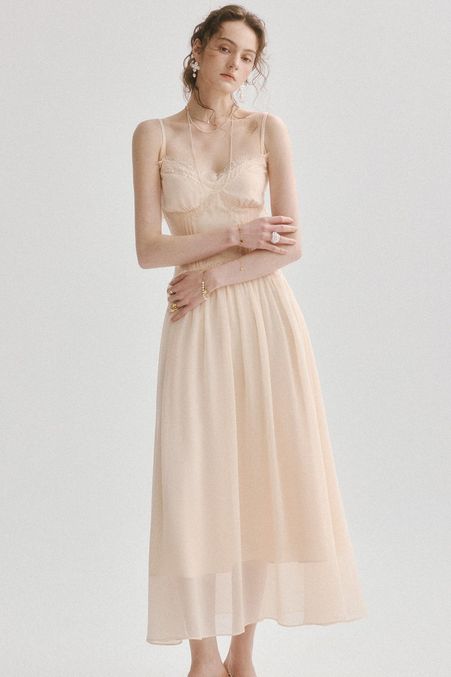 A woman modeling a sleeveless midi dress in a soft blush beige color, featuring a structured lace bodice and a flowing chiffon skirt, set against a plain light gray studio background.