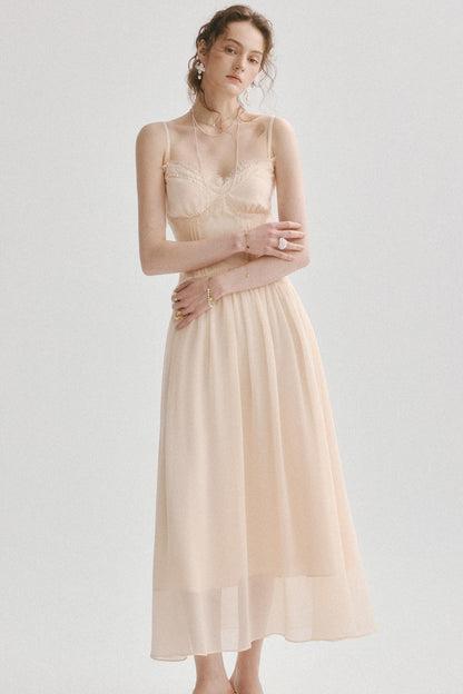 A woman modeling a sleeveless midi dress in a soft blush beige color, featuring a structured lace bodice and a flowing chiffon skirt, set against a plain light gray studio background.