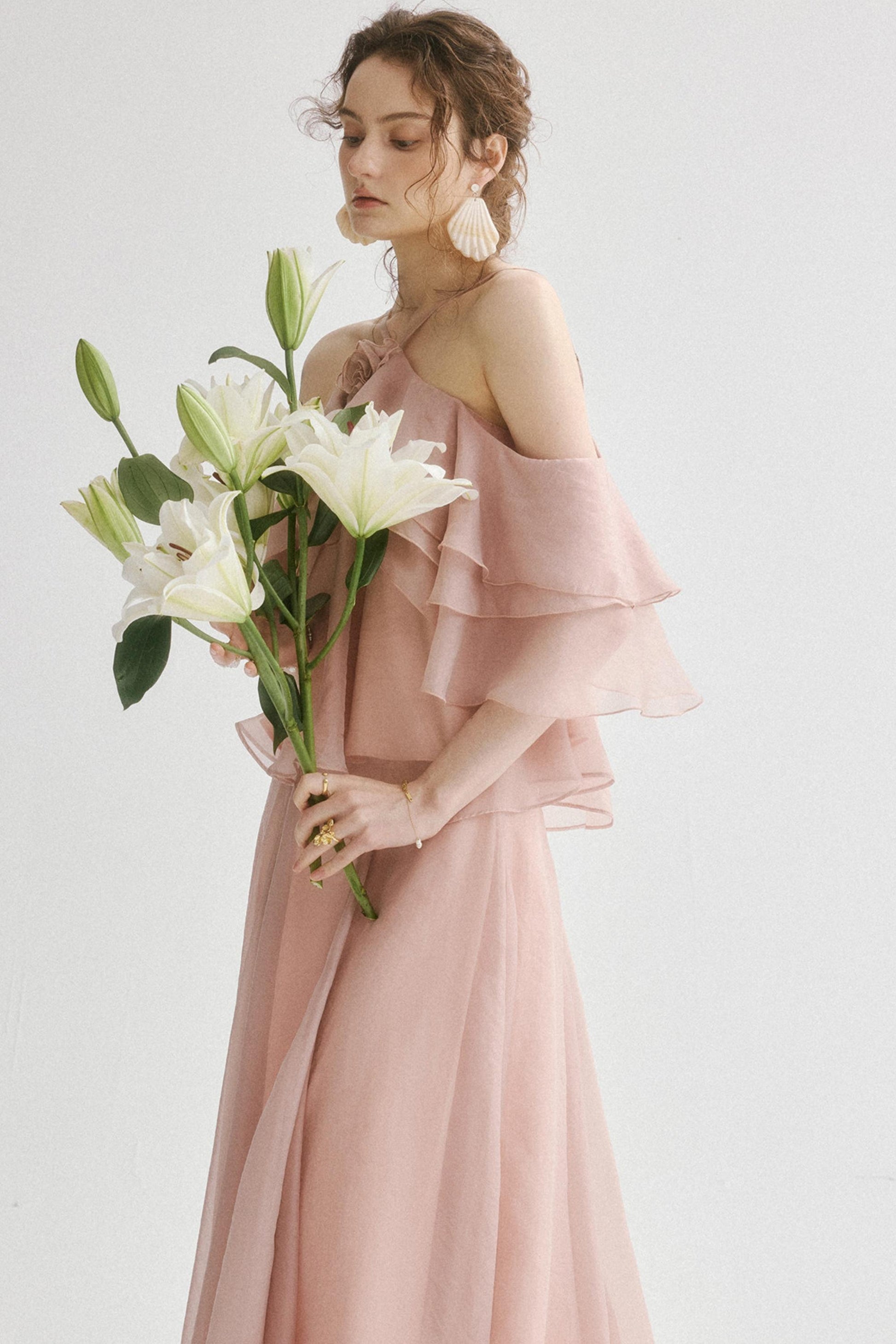 A woman wearing a dusty pink off-the-shoulder tiered gown and holding a bouquet of white lilies, set against a plain light gray background.