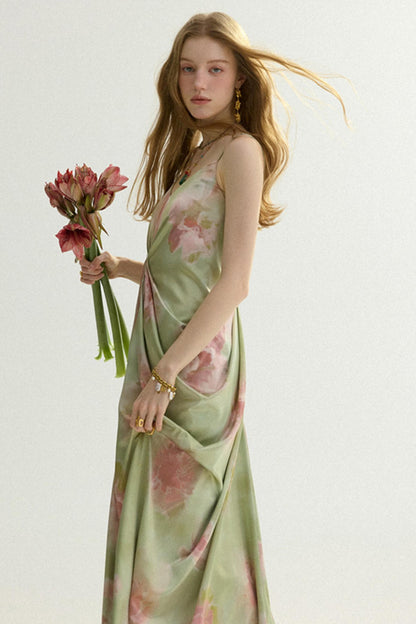 A woman wearing a sleeveless sage green satin maxi dress with a pink floral print, holding a bouquet of pink flowers against a plain white studio background.