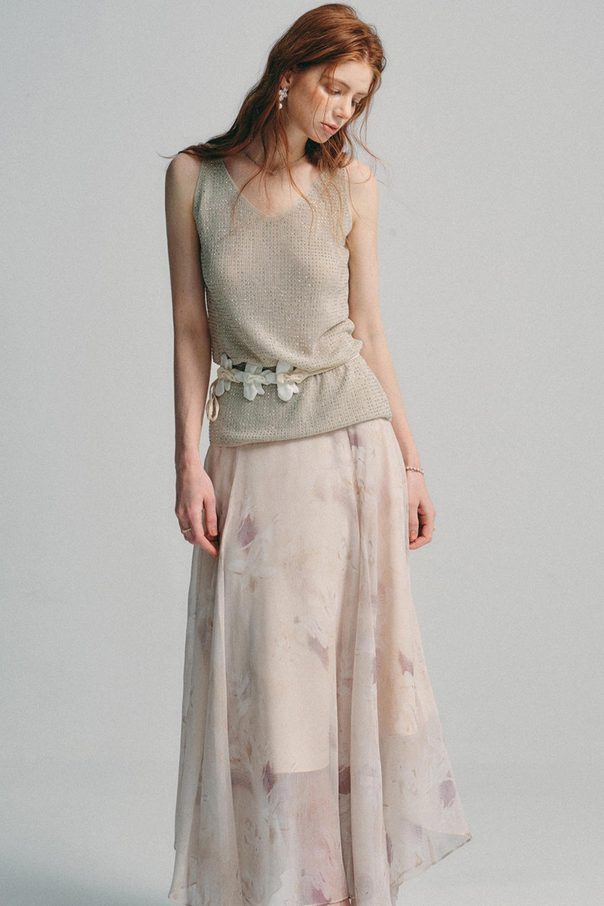 A woman modeling a sleeveless textured knit top in a light beige/taupe color paired with a long, flowing sheer skirt featuring a soft pink and beige floral pattern, set against a plain light gray studio background.