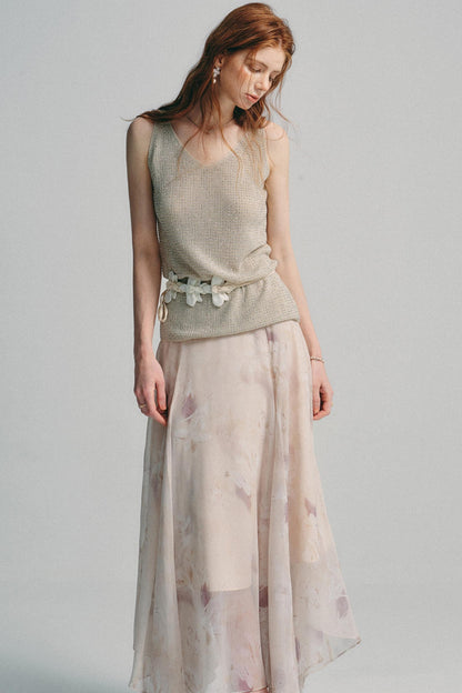 A woman modeling a sleeveless textured knit top in a light beige/taupe color paired with a long, flowing sheer skirt featuring a soft pink and beige floral pattern, set against a plain light gray studio background.