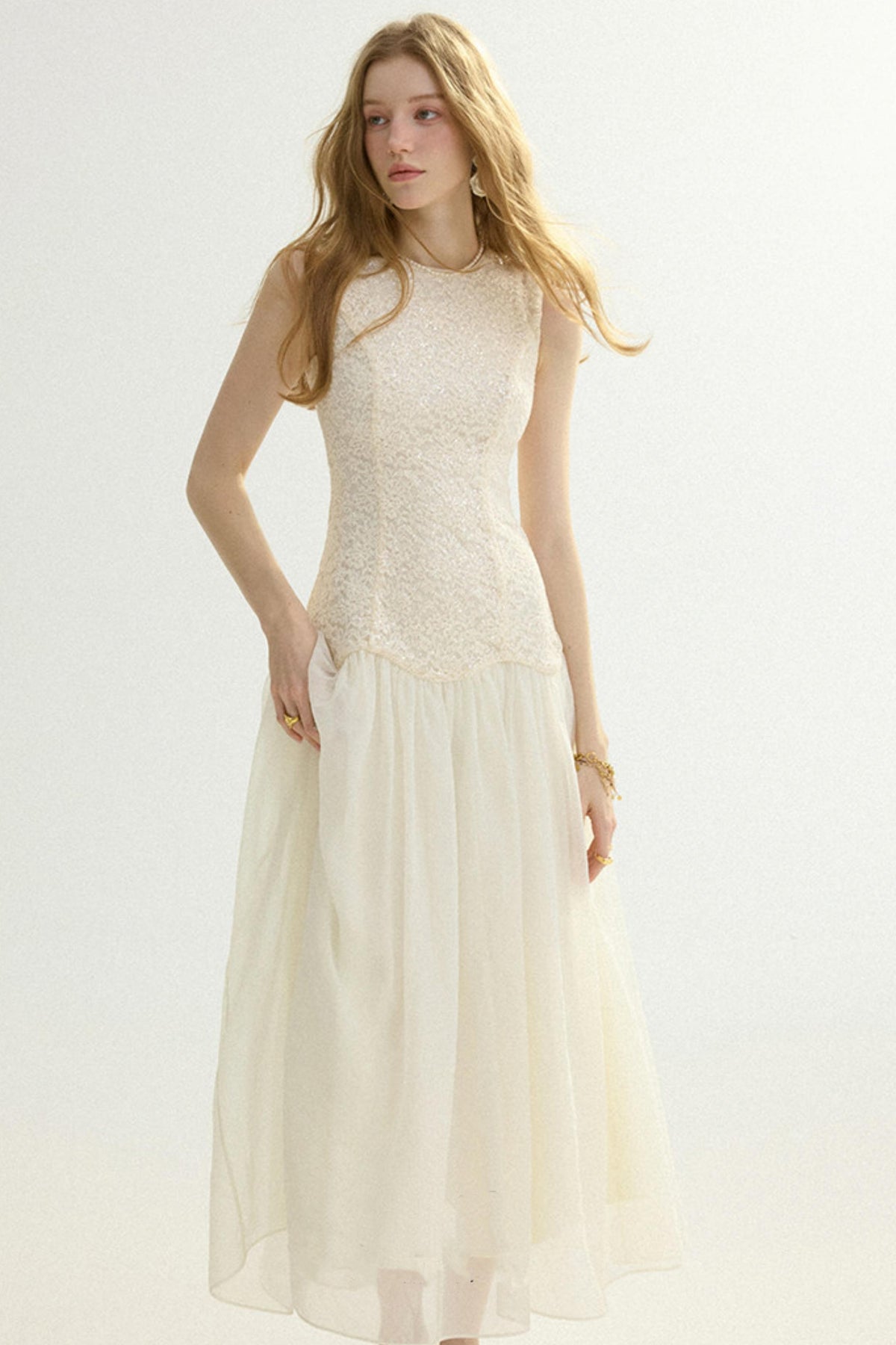 A woman modeling a sleeveless cream maxi dress with a textured lace bodice and a flowing tulle skirt, set against a plain light background.