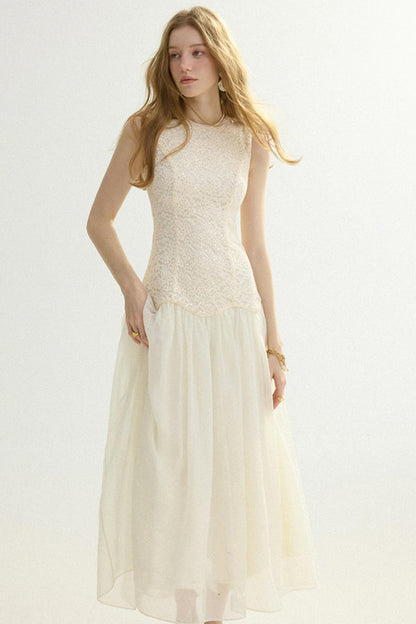 A woman modeling a sleeveless cream maxi dress with a textured lace bodice and a flowing tulle skirt, set against a plain light background.