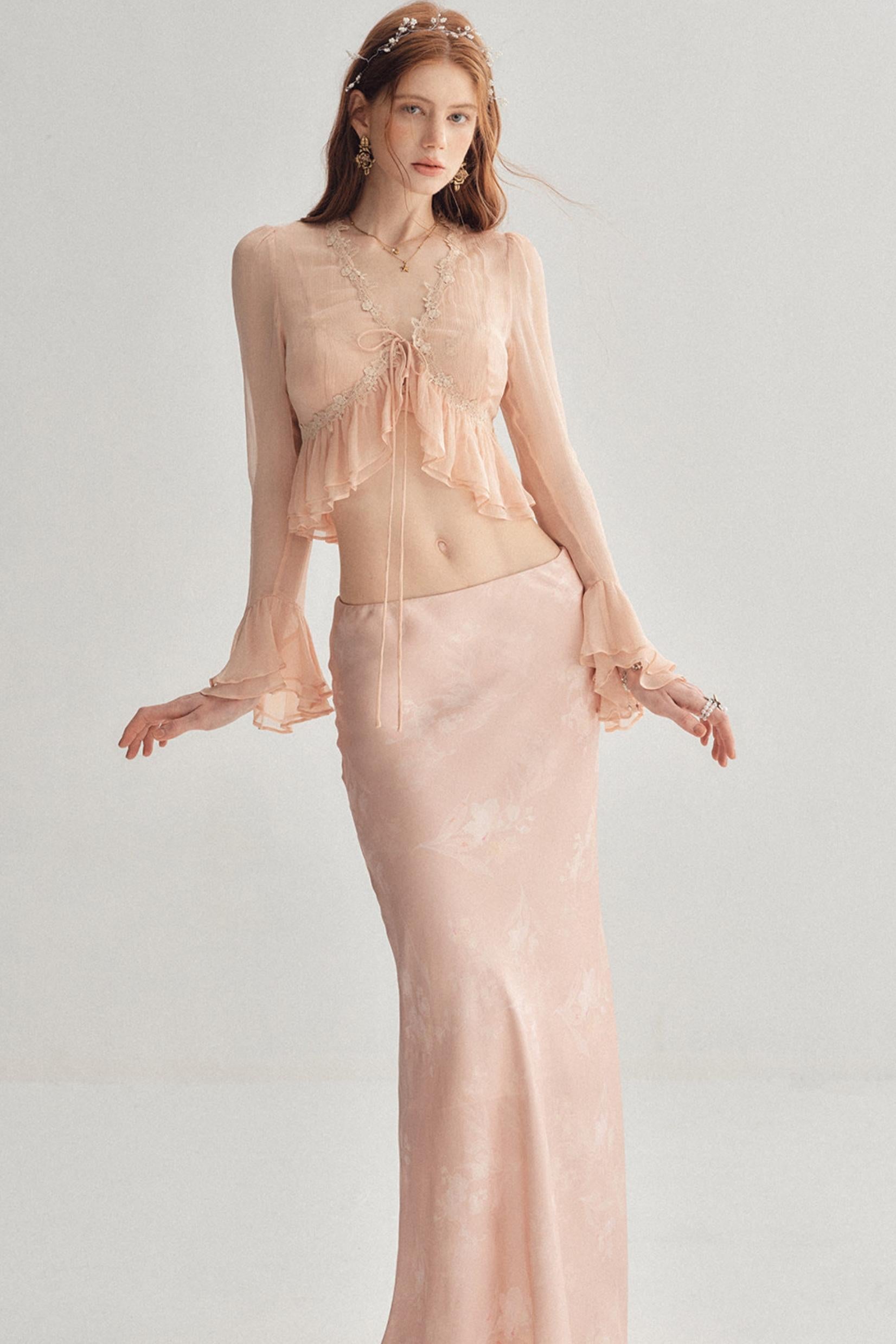 A woman modeling a two-piece formal outfit in a soft blush pink color. The set includes a sheer long-sleeve crop top with ruffle details and a matching satin-like floor-length skirt, set against a plain light gray studio background.