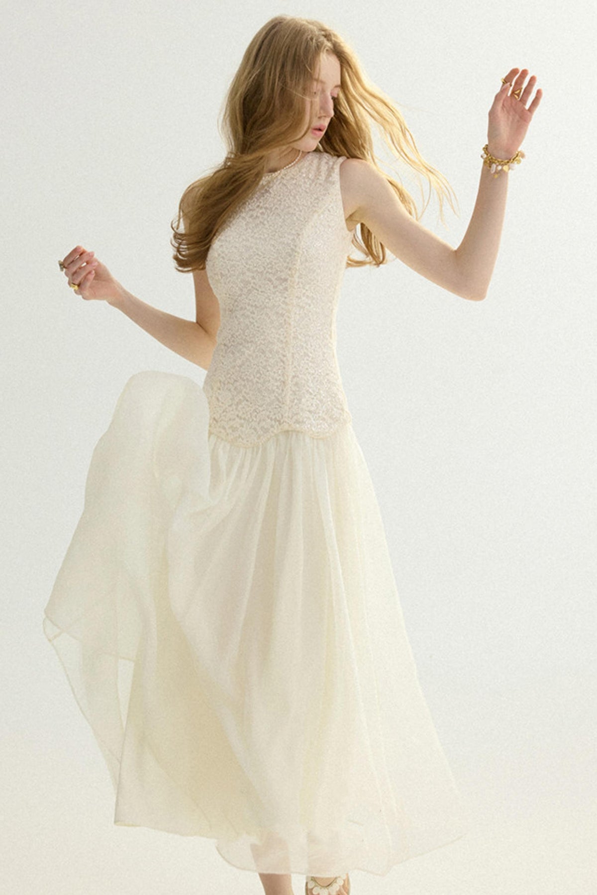 A woman wearing a sleeveless cream lace bodice with a flowing tulle skirt, posed against a plain light background.