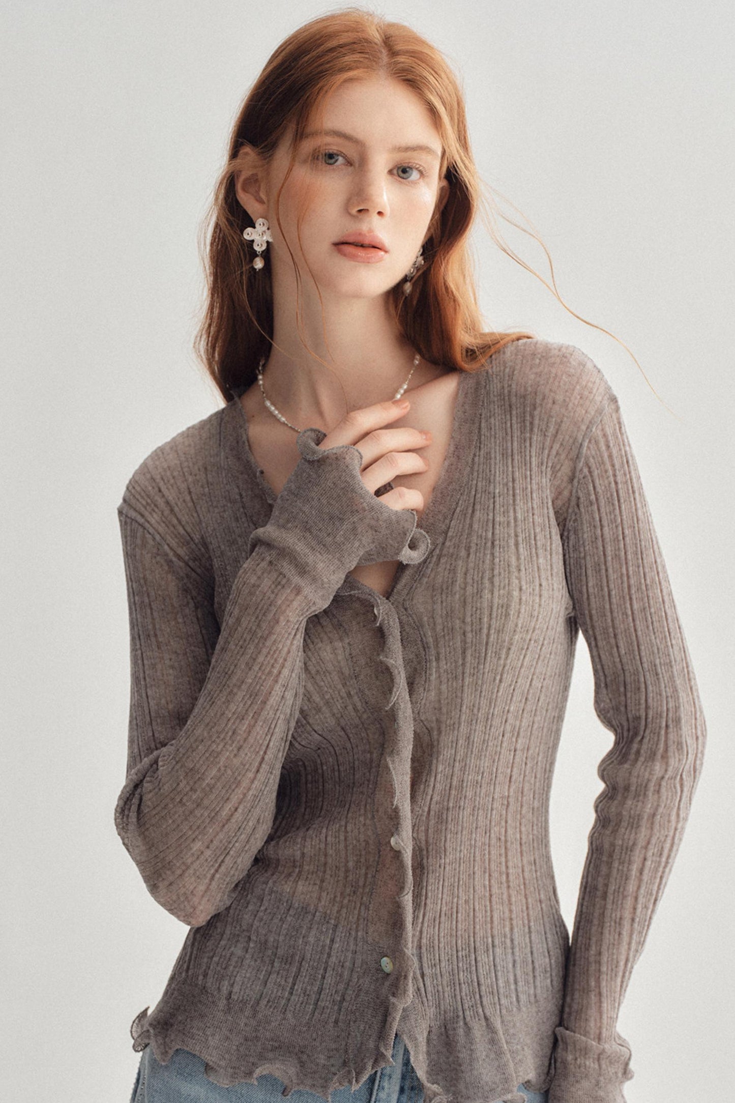 A woman wearing a ribbed knit cardigan in a taupe-brown color, styled with light blue jeans and pearl jewelry against a plain light gray background.