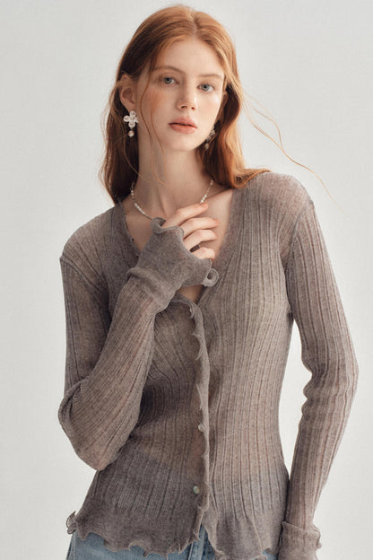 A woman wearing a ribbed knit cardigan in a taupe-brown color, styled with light blue jeans and pearl jewelry against a plain light gray background.