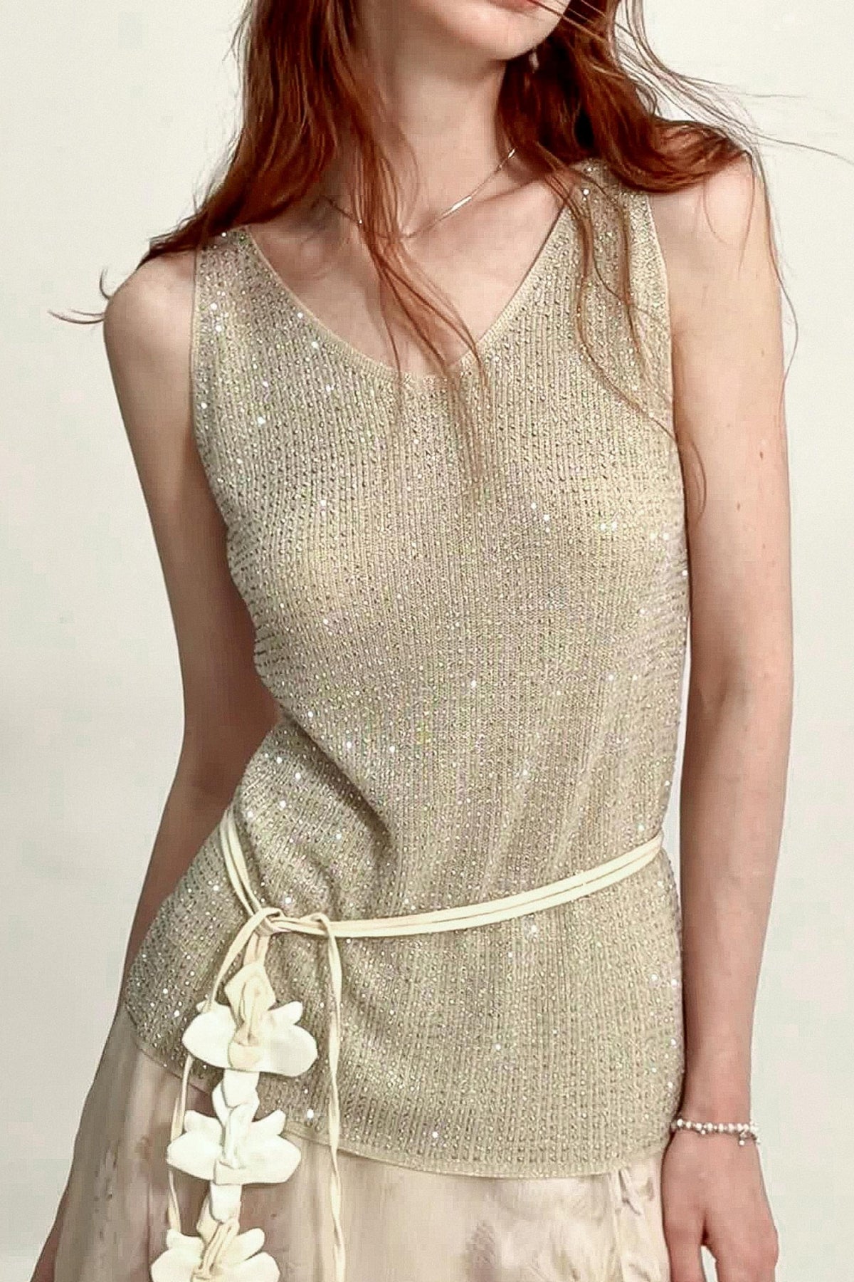 A woman wearing a sleeveless shimmering knit top in a light champagne beige color, paired with a matching beige skirt and white flower accents at the waist.