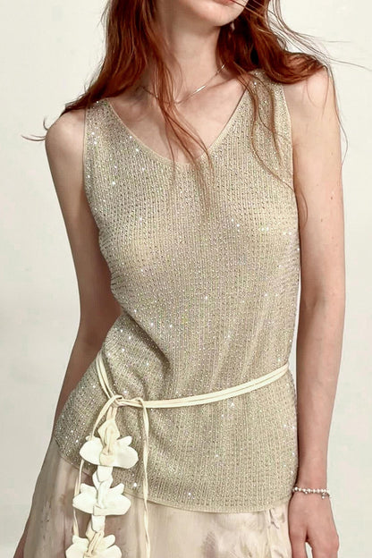 A woman wearing a sleeveless shimmering knit top in a light champagne beige color, paired with a matching beige skirt and white flower accents at the waist.