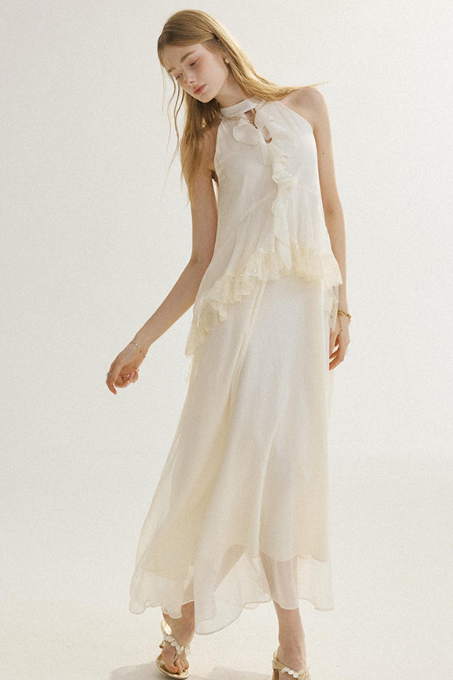 A woman modeling a sleeveless, high-neck, floor-length cream dress with layered ruffle details and a sheer overlay, paired with metallic gold sandals against a plain white studio background.