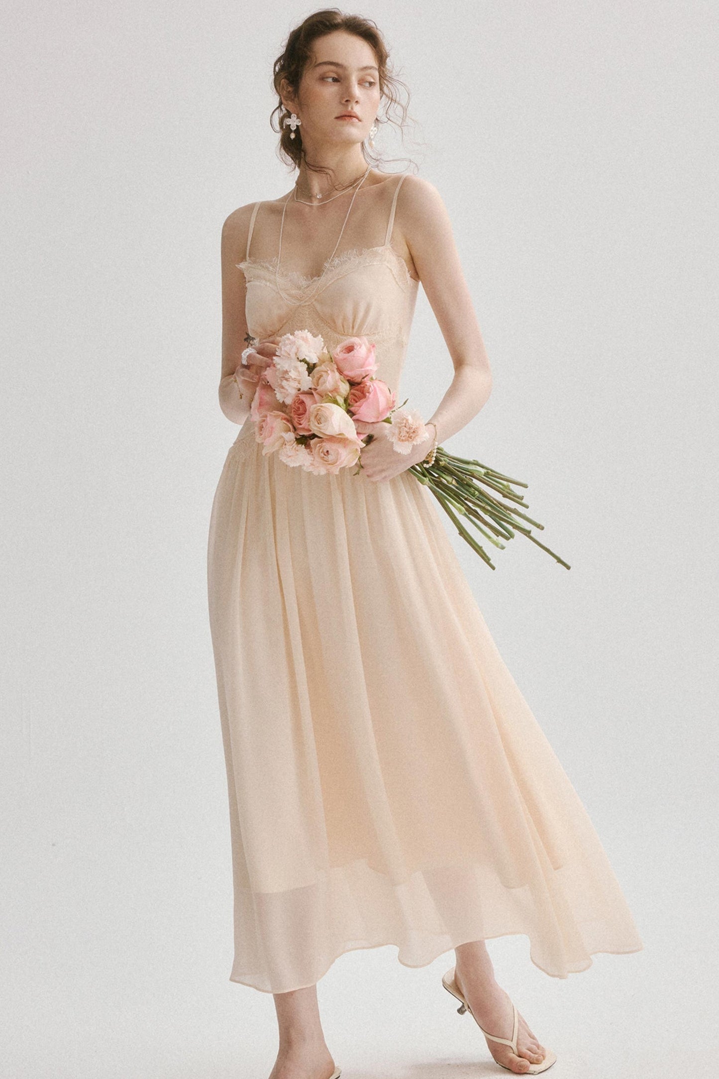A woman modeling a sleeveless, flowing midi dress in a soft blush pink or light peach tone, holding a bouquet of pink flowers against a plain light gray studio background.