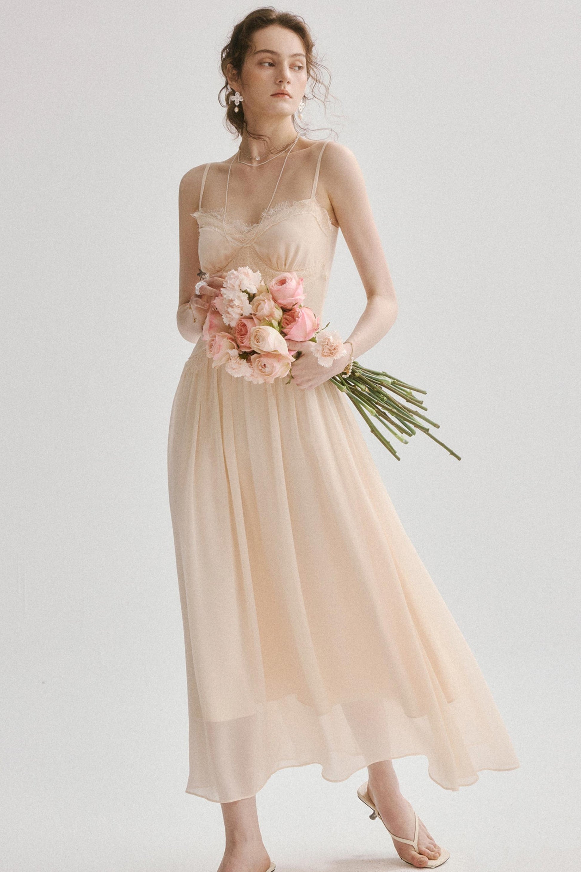 A woman modeling a sleeveless, flowing midi dress in a soft blush pink or light peach tone, holding a bouquet of pink flowers against a plain light gray studio background.