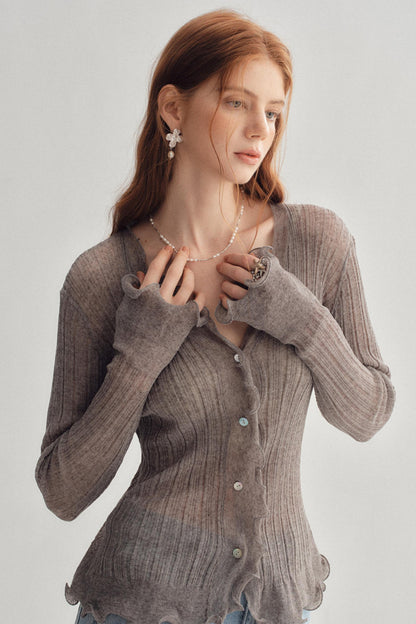A woman wearing a ribbed knit long-sleeve cardigan in taupe gray, styled with a delicate pearl necklace and earrings against a light gray studio background.