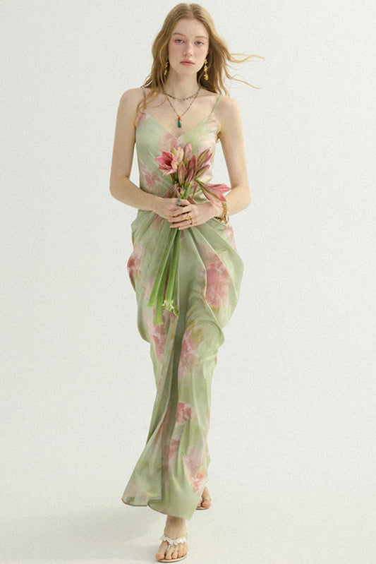 A woman modeling a light sage green maxi dress with a pink and green floral print, holding a bouquet of flowers against a plain white studio background.