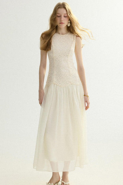 A woman modeling a sleeveless cream maxi dress with a textured lace bodice and a flowing tulle skirt, set against a plain light background.