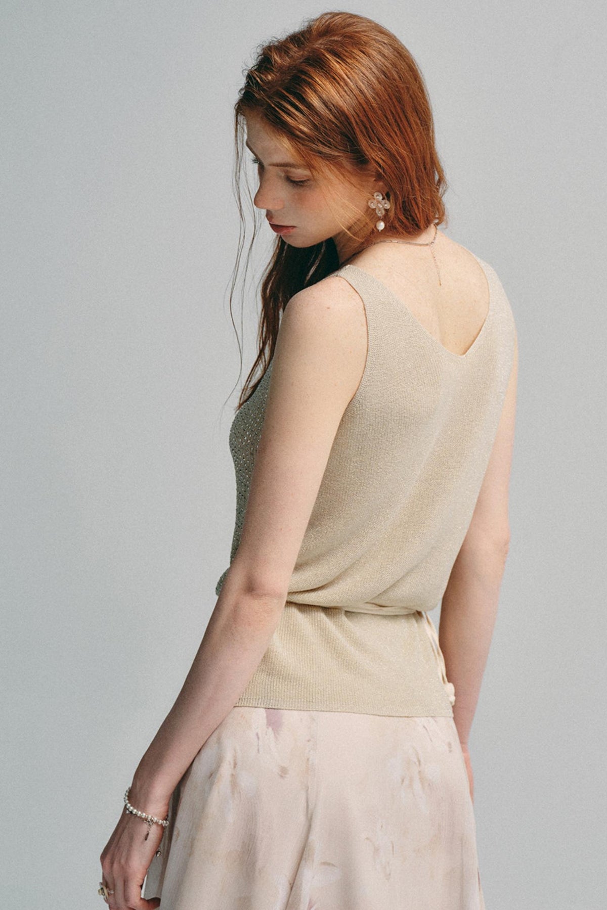 A woman with red hair is shown from the back wearing a beige sleeveless knit top and a light pink skirt against a solid gray background.