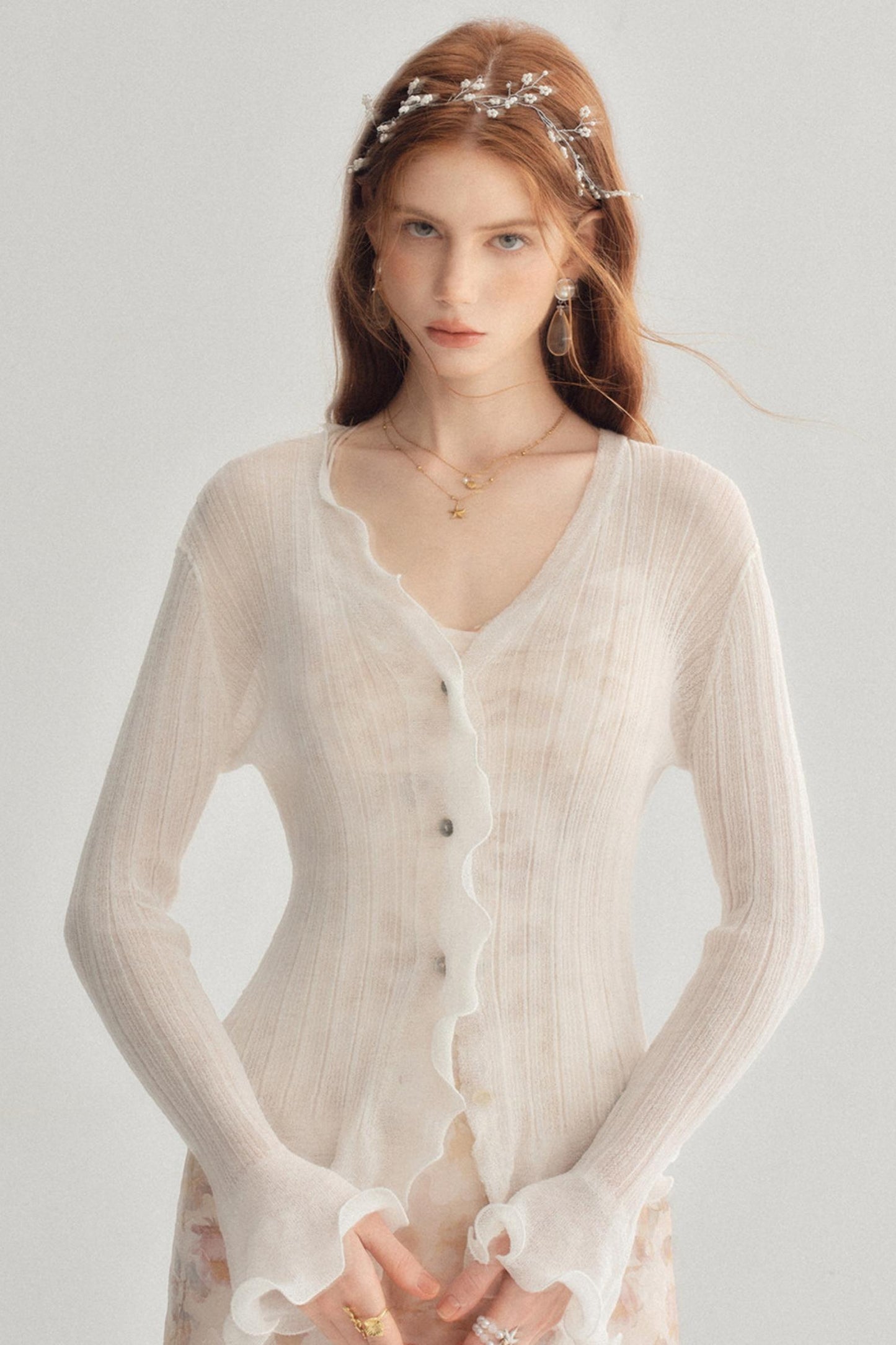 A woman wearing a sheer, long-sleeve cream cardigan with scalloped edges and button details, styled with a pearl headband and gold jewelry against a light gray background.