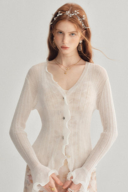 A woman wearing a sheer, long-sleeve cream cardigan with scalloped edges and button details, styled with a pearl headband and gold jewelry against a light gray background.