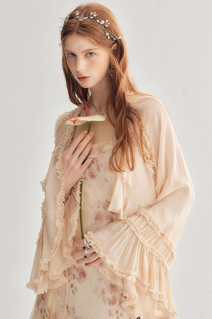 A woman with long red hair wears a light peach-beige floral robe with ruffled trim over a matching floral dress. She is posed against a plain light gray background, holding a flower stem near her chest.