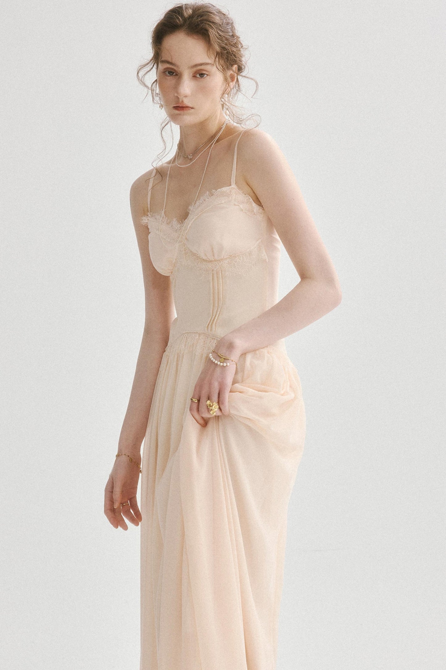 A woman modeling a pale blush-beige maxi dress with thin straps, a ruffled bust, and a gathered skirt, photographed against a clean light gray studio background.