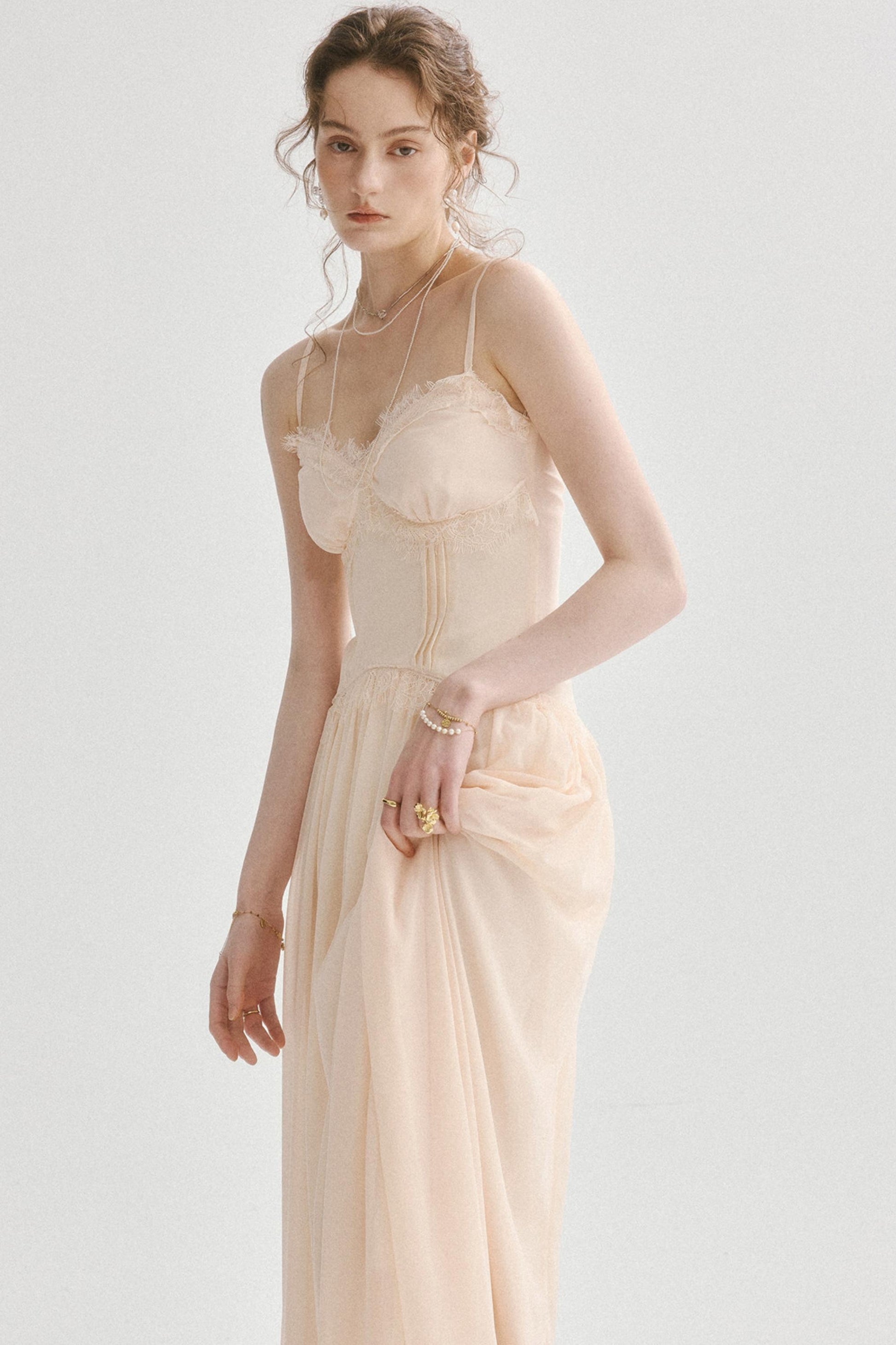 A woman modeling a pale blush-beige maxi dress with thin straps, a ruffled bust, and a gathered skirt, photographed against a clean light gray studio background.