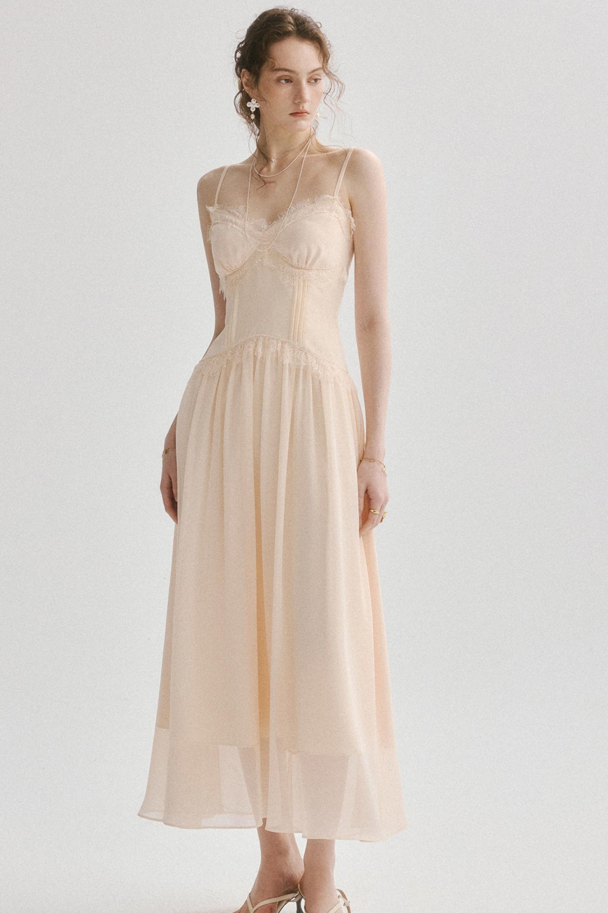 A woman modeling a pale blush-beige spaghetti-strap maxi dress with a structured corset-style bodice and flowing sheer skirt, photographed against a light gray studio background.