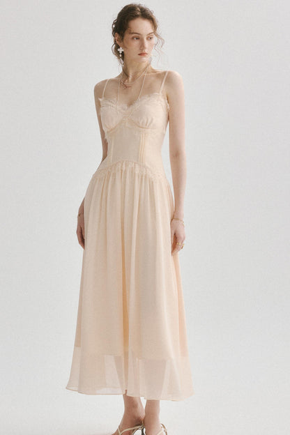 A woman modeling a pale blush-beige spaghetti-strap maxi dress with a structured corset-style bodice and flowing sheer skirt, photographed against a light gray studio background.