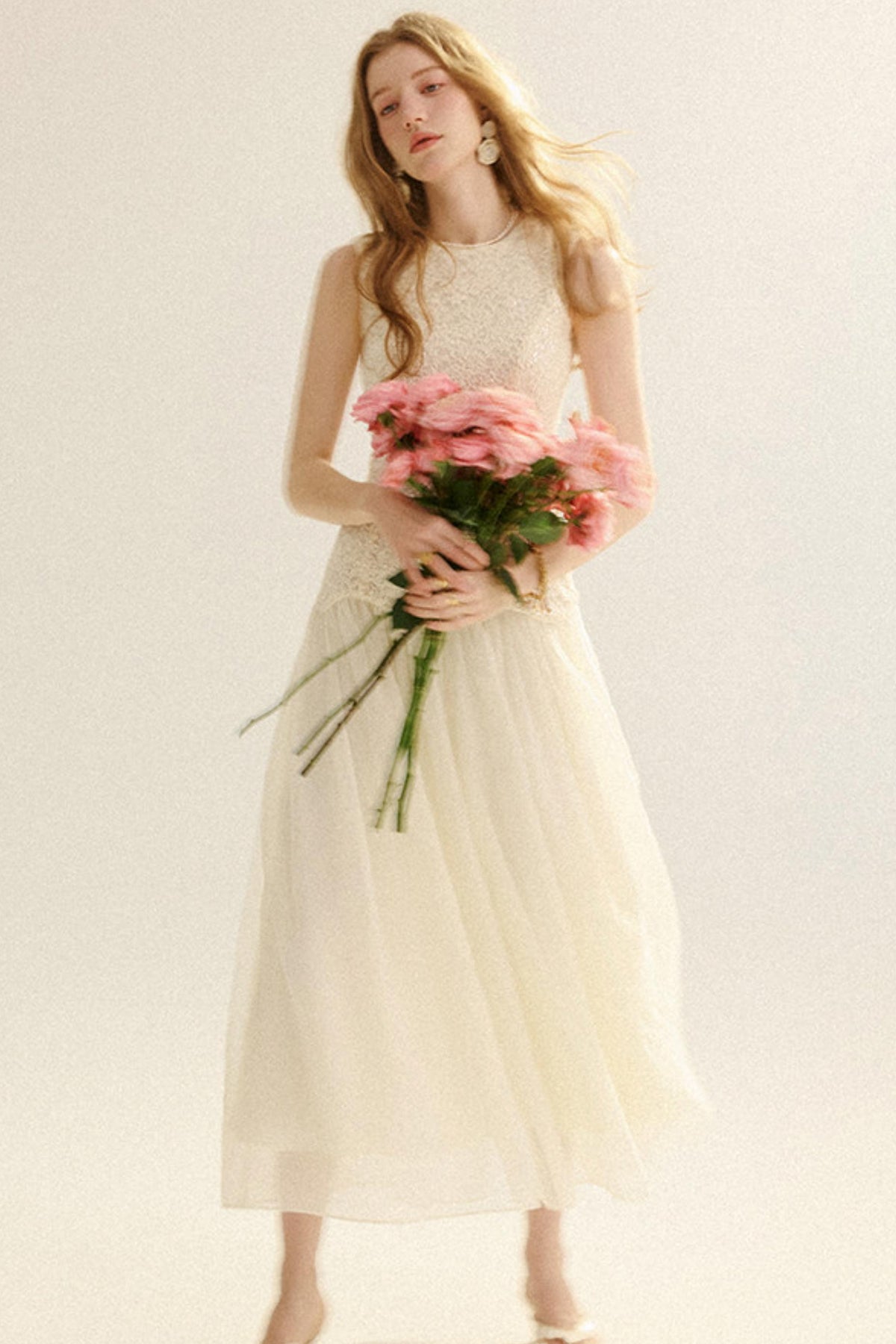 A woman wearing a sleeveless cream tulle midi dress with a lace bodice, holding a bouquet of pink flowers against a plain light background.
