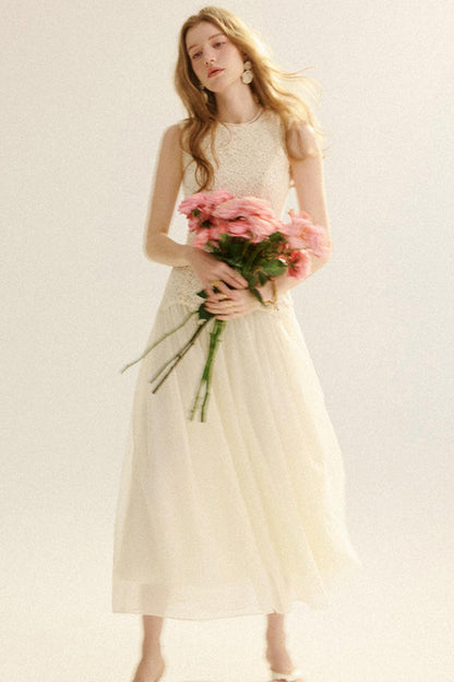 A woman wearing a sleeveless cream tulle midi dress with a lace bodice, holding a bouquet of pink flowers against a plain light background.