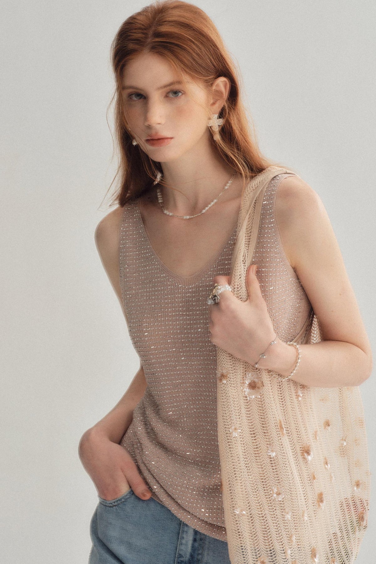 A woman wearing a shimmering beige sleeveless knit top and blue jeans, holding a matching beige beaded shoulder bag against a plain light gray background.